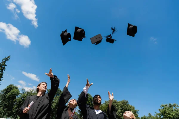 Low angle view of multiethnic bachelors in gowns throwing caps outdoors - foto de stock