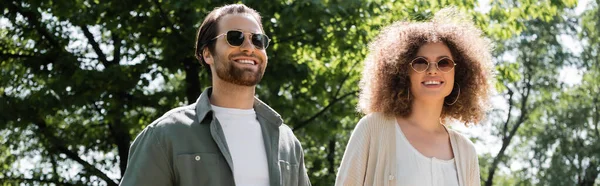 Femme bouclée et homme heureux dans des lunettes de soleil élégantes marchant dans le parc, bannière — Photo de stock