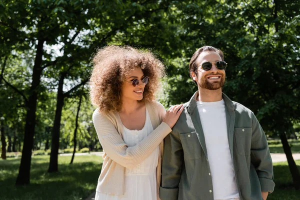 Mulher encaracolado e homem feliz em óculos de sol elegantes andando no parque — Fotografia de Stock