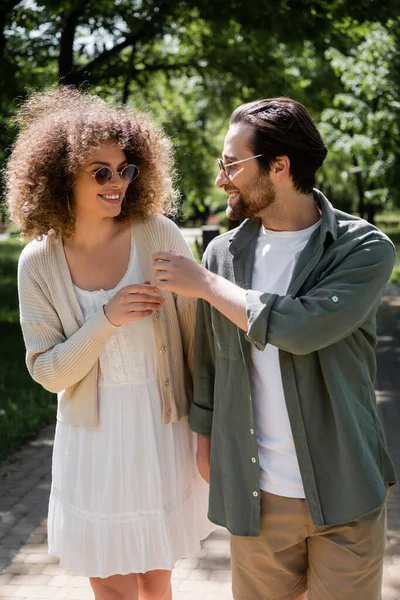 Femme bouclée et homme joyeux dans des lunettes de soleil élégantes marchant dans le parc — Photo de stock