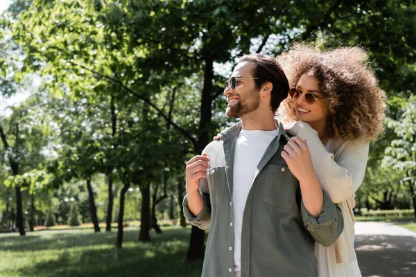 Mulher encaracolado abraçando homem feliz em óculos de sol elegantes no parque — Fotografia de Stock