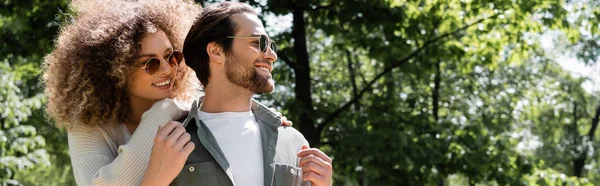 Кудрявая женщина обнимает счастливого мужчину в стильных солнечных очках в парке, баннер — стоковое фото