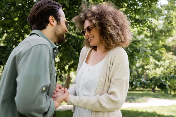 Curly woman holding hands with happy man in stylish sunglasses in park — Foto stock