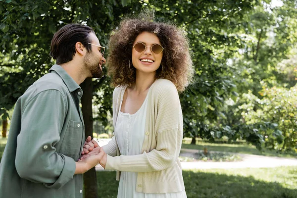 Весела жінка тримає руки з щасливим чоловіком в стильних сонцезахисних окулярах в парку — стокове фото