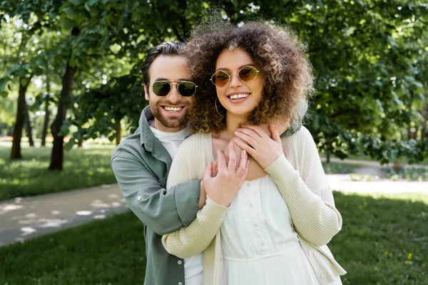 Счастливый мужчина в стильных солнцезащитных очках обнимает кудрявую девушку в парке — стоковое фото