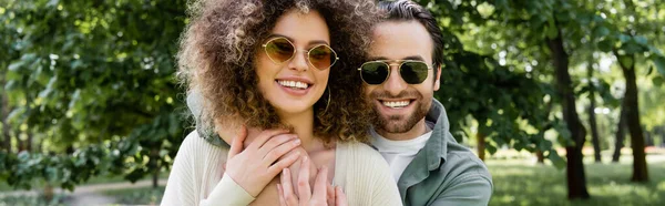 Homme heureux dans des lunettes de soleil élégantes étreignant petite amie bouclée dans le parc, bannière — Photo de stock