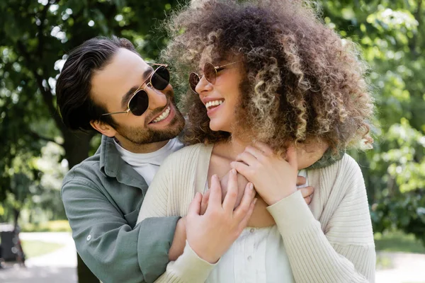 Happy man in stylish sunglasses hugging pleased girlfriend in park - foto de stock