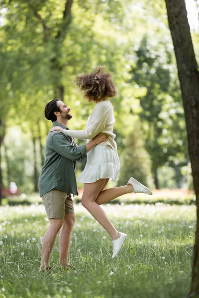 Pleine longueur de jeune homme heureux soulevant petite amie bouclée en robe d'été dans le parc vert — Photo de stock