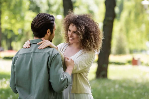 Happy and curly woman in cardigan hugging boyfriend in green park — Fotografia de Stock