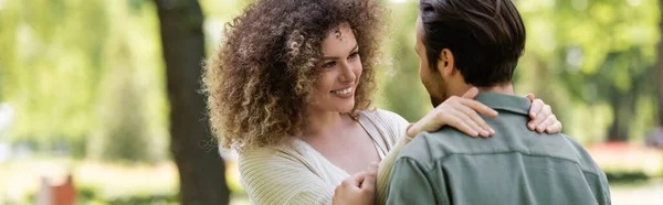 Счастливая и кудрявая женщина в кардигане обнимает бойфренда в зеленом парке, баннер — стоковое фото