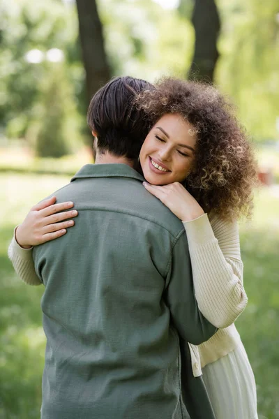 Cheerful and curly woman in cardigan hugging boyfriend in green park — Stock Photo