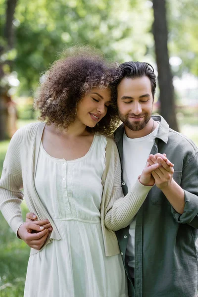 Happy and curly woman in cardigan holding hands with boyfriend in green park — Stock Photo