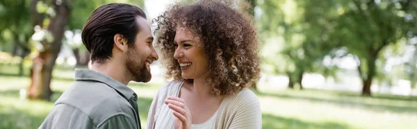 Mujer feliz y rizado en cárdigan mirando alegre novio en verde parque, pancarta - foto de stock