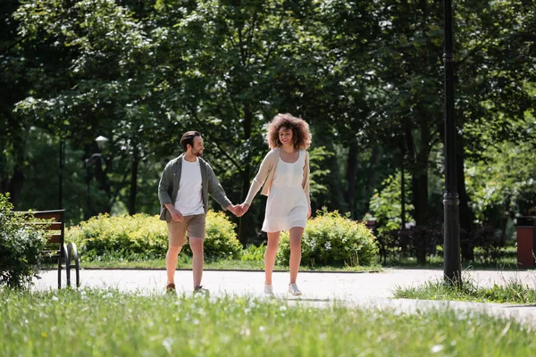 Повна довжина щасливої молодої пари тримає руки під час прогулянки разом в літньому парку — стокове фото