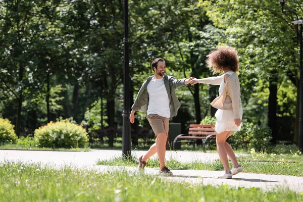 Full length of cheerful young couple holding hands while walking together in summer park — Stockfoto