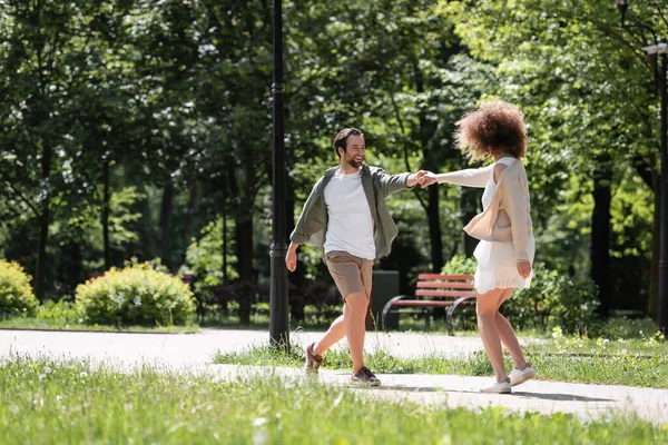 Full length of cheerful young couple holding hands while walking together in summer park - foto de stock