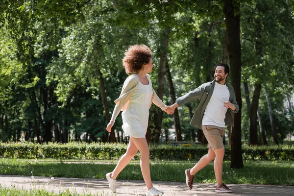 Longitud completa de pareja joven positiva tomados de la mano mientras corren juntos en el parque de verano - foto de stock