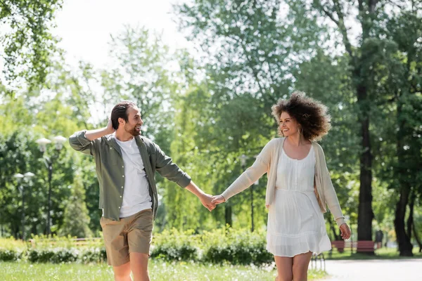 Pleased young couple holding hands while walking together in summer park — Stock Photo