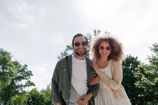 Uomo allegro e felice donna riccia in occhiali da sole alla moda sorridente nel parco — Foto stock