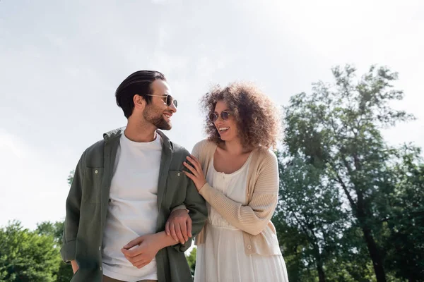 Homem alegre e mulher encaracolada feliz em óculos de sol na moda sorrindo e olhando um para o outro no parque — Fotografia de Stock