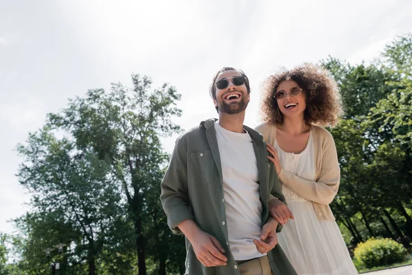 Homem alegre e mulher encaracolada feliz em óculos de sol na moda sorrindo no parque — Fotografia de Stock