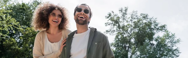 Hombre alegre y mujer rizada positiva en gafas de sol de moda sonriendo en el parque, pancarta - foto de stock