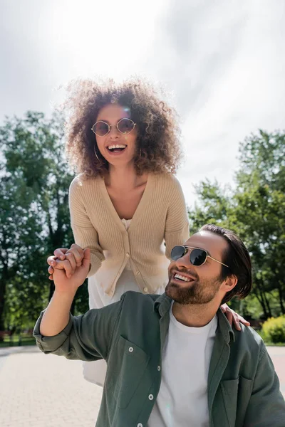 Веселый мужчина и счастливая кудрявая женщина в модных солнцезащитных очках, держась за руки и улыбаясь в парке — стоковое фото