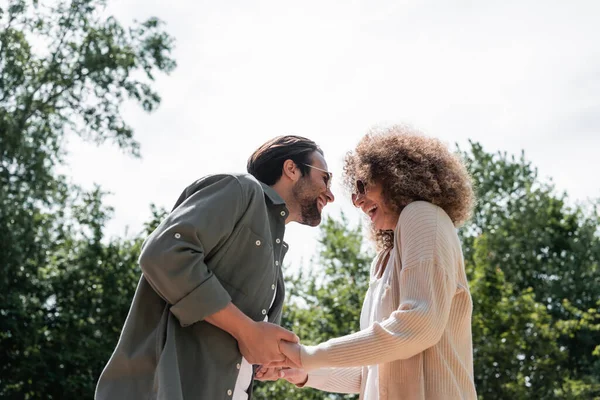 Tiefansicht eines fröhlichen Mannes und einer glücklichen lockigen Frau mit trendiger Sonnenbrille, die sich im Park an den Händen halten — Stockfoto