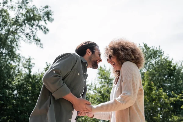 Low angle view of cheerful man and happy curly woman in trendy sunglasses holding hands in park — Fotografia de Stock