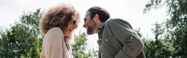 Low angle view of cheerful man and happy curly woman in trendy sunglasses looking at each other in park, banner — Photo de stock