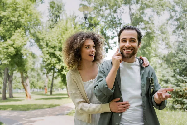 Lockige Frau umarmt ihren glücklichen Freund, der im grünen Park mit dem Smartphone spricht — Stockfoto