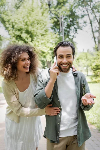 Curly woman hugging cheerful boyfriend talking on smartphone in green park — Stockfoto