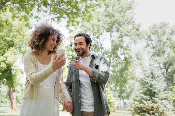 Homem feliz apontando para smartphone perto de namorada encaracolado no parque verde — Fotografia de Stock