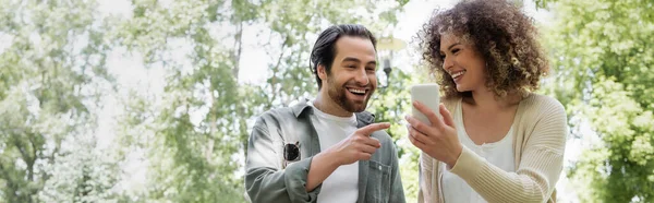 Heureux homme pointant vers smartphone près de petite amie bouclée dans le parc vert, bannière — Photo de stock