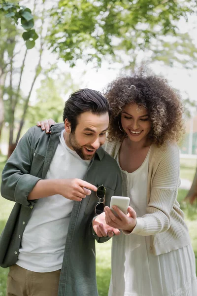 Hombre excitado señalando con el dedo en el teléfono inteligente cerca de novia rizada en el parque verde - foto de stock