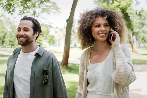 Heureuse femme bouclée parler sur smartphone près petit ami souriant dans le parc — Photo de stock