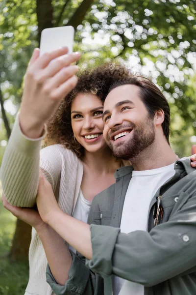 Joyeux jeune couple prenant selfie sur smartphone flou dans le parc d'été vert — Photo de stock