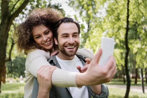 Щаслива молода пара сідає на розмитий смартфон у літньому парку. — стокове фото