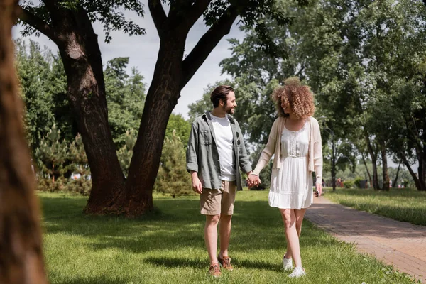 Full length of happy young couple holding hands while standing on grass in summer park — Fotografia de Stock