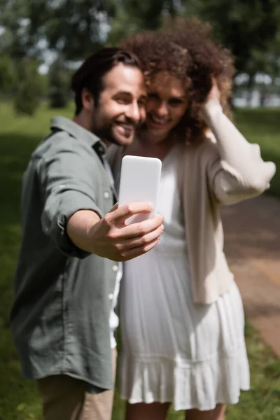 Pareja borrosa positiva tomando selfie en el teléfono inteligente en el parque de verano - foto de stock