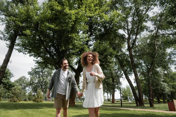 Cheerful man and pleased curly woman in dress holding hands in summer park — Stockfoto