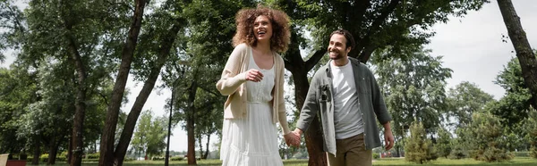 Cheerful man and pleased curly woman in dress holding hands in summer park, banner — Fotografia de Stock