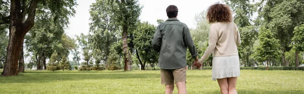 Vue arrière de la femme bouclée en robe et de l'homme en vêtements d'été marchant ensemble dans le parc, bannière — Photo de stock