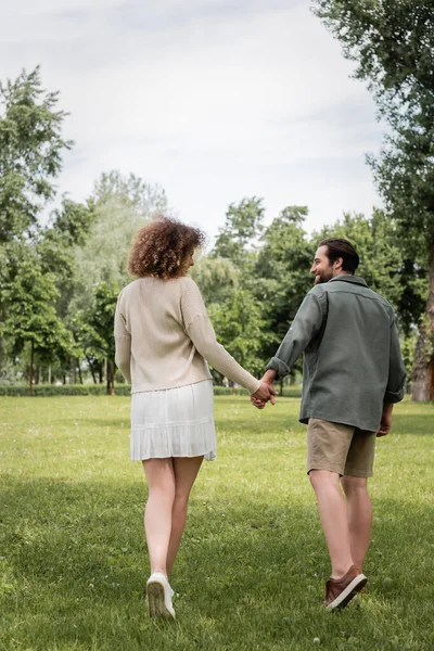 Vista posteriore della donna riccia in abito e l'uomo in vestiti estivi che si tiene per mano nel parco — Foto stock