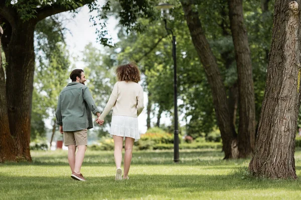Вид в полный рост кудрявой женщины в платье и мужчины в летней одежде, держащегося за руки в парке — стоковое фото