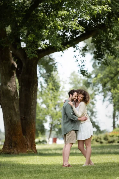 Full length of cheerful couple in summer clothes hugging in park — Stock Photo