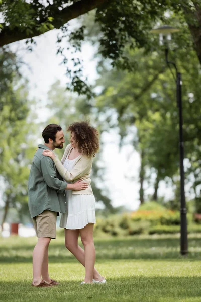 Toute la longueur du couple souriant en vêtements d'été étreignant dans le parc — Photo de stock