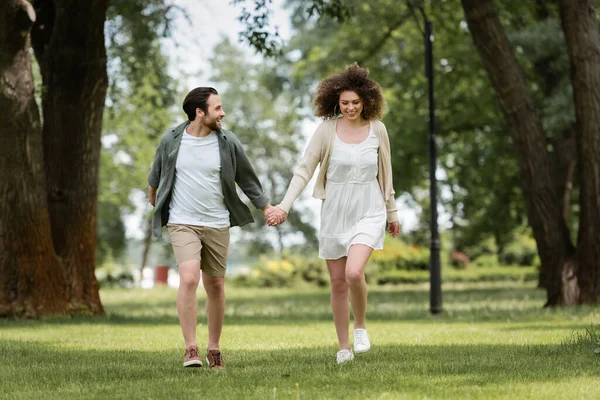 Full length of smiling couple in summer clothes holding hands and walking in park — Stock Photo