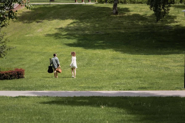 Longitud completa del hombre en ropa de verano llevando la guitarra y cesta de mimbre picnic cerca de la novia en el parque verde - foto de stock