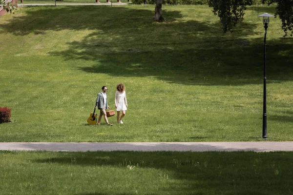 Full length of man in summer clothes carrying guitar and picnic wicker basket near happy girlfriend in green park — Stockfoto